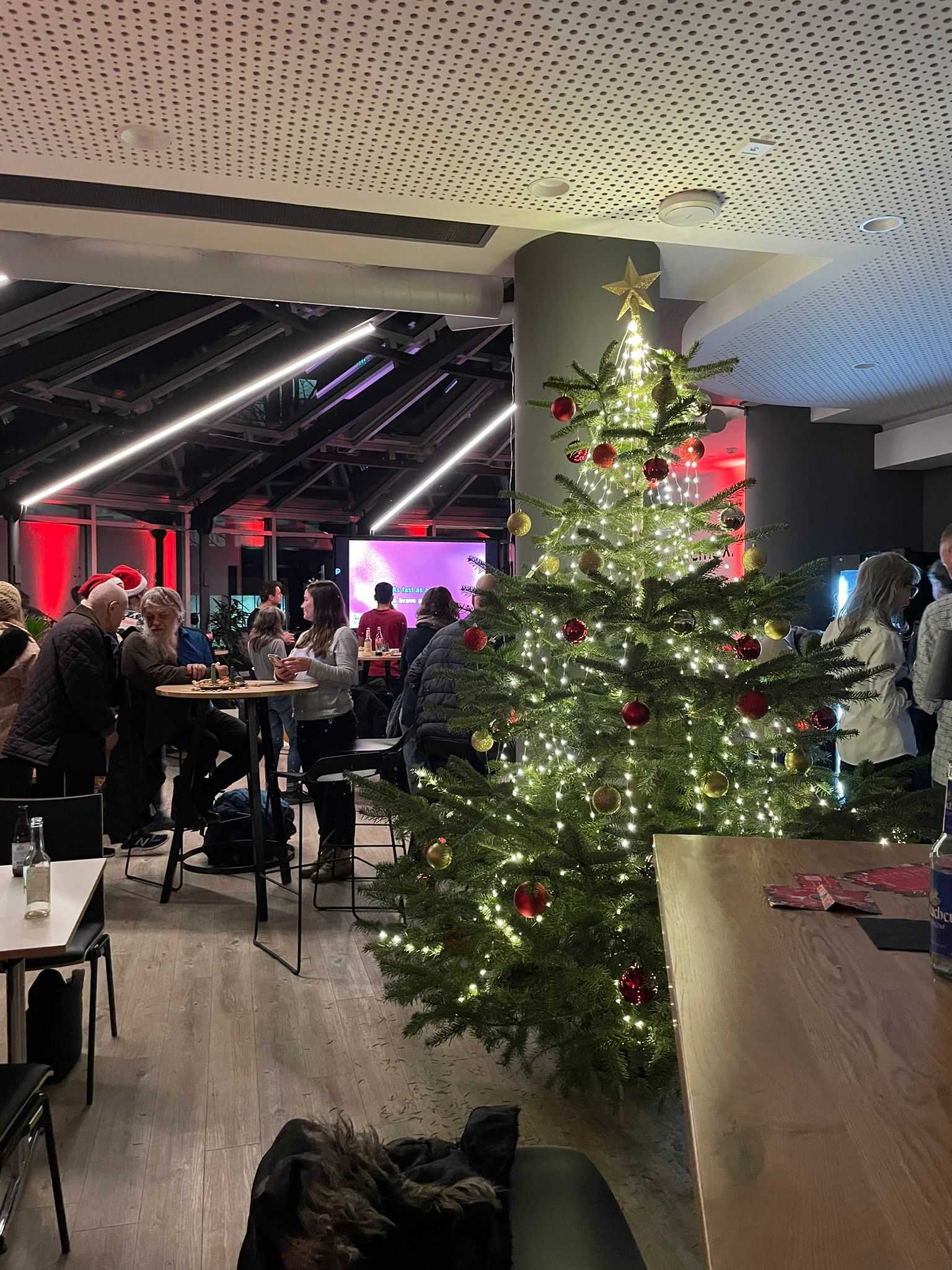 Gäste im Gespräch an Stehtischen rund um den Weihnachtsbaum, im Hintergrund läuft Christmas-Karaoke
