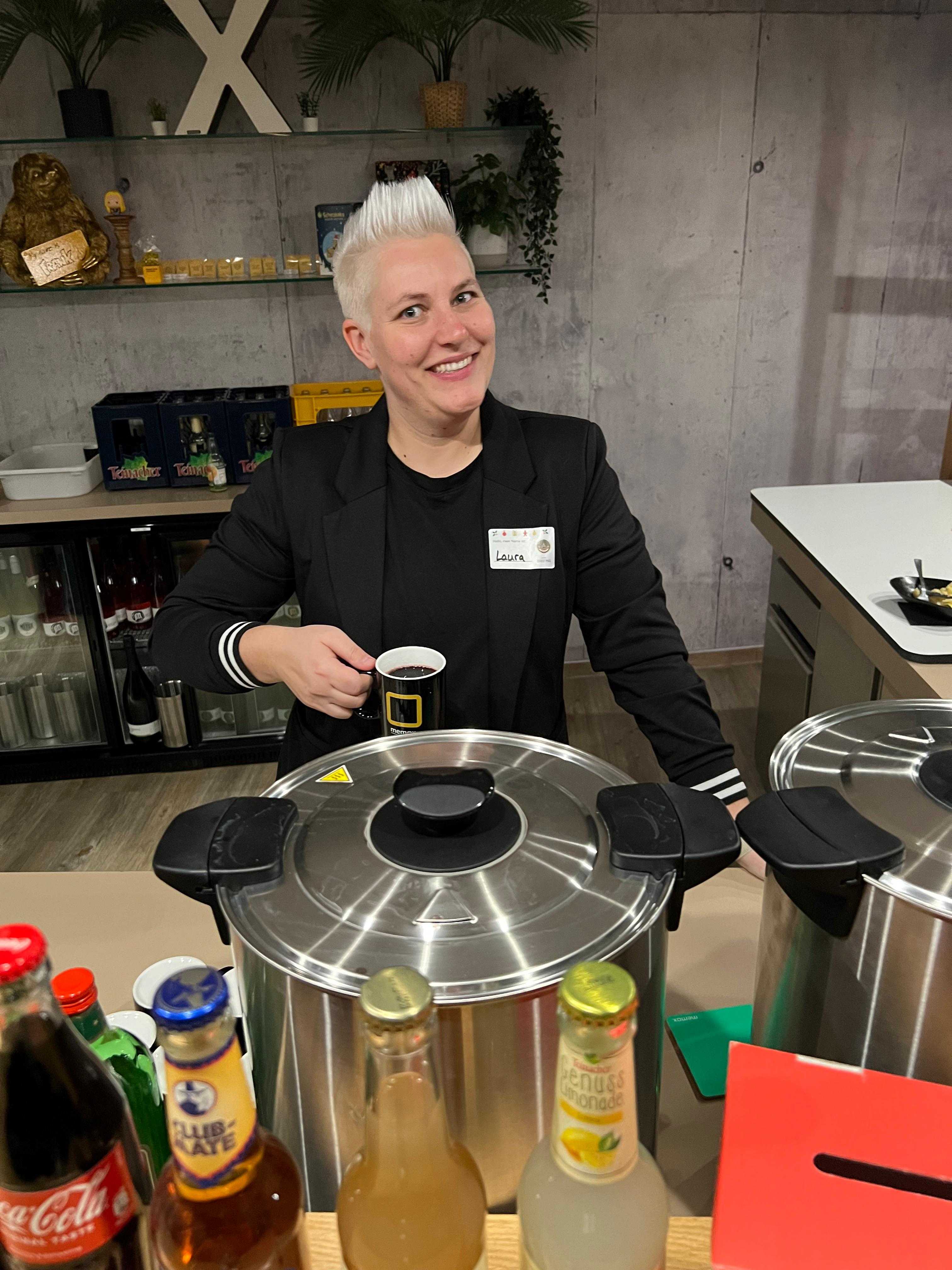 Lächelnde Helferin mit Tasse hinter den großen Kesseln der Glühwein- und Punschstation
